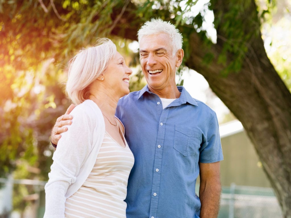 Older Couple Smiling In Front Of Trees With Sun Shining Through Older Couple Smiling In Front Of Trees With Sun Shining Through